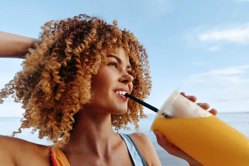 Smiling woman with curly hair drinks a frozen orange beverage through a straw on a sunny bright beach, ocean horizon behind her, summer refreshment and vacation relaxation outdoors