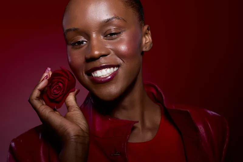 Close-up portrait of happy black woman with dark skin holding red rose near face, smiling with closed eyes. Studio shot with red background and stylish red outfit. Concept of beauty and elegance.