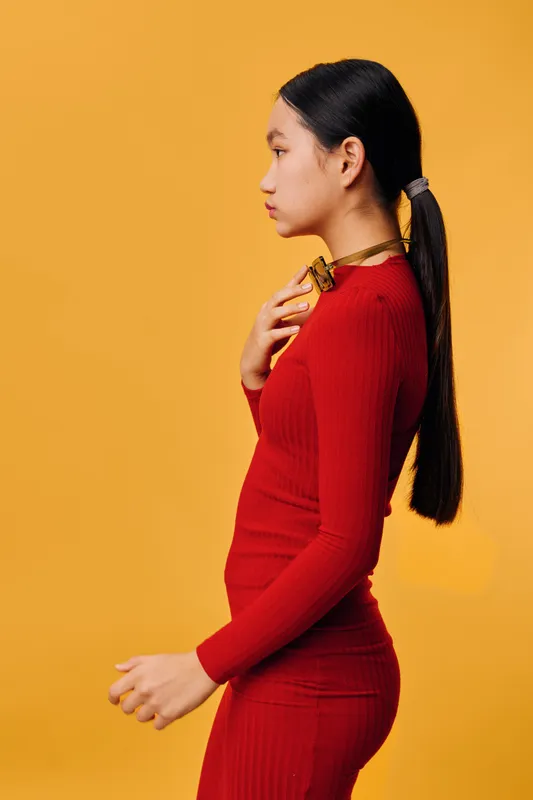 Woman in red dress with long black hair tied in ponytail standing in profile on yellow background. Fashion, style and portrait of young female model with elegant pose and casual look.