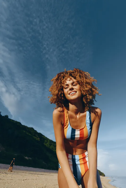 Young woman with curly hair wearing colorful striped swimsuit smiling outdoors on sunny beach with blue sky and green hills in background relaxed and happy mood
