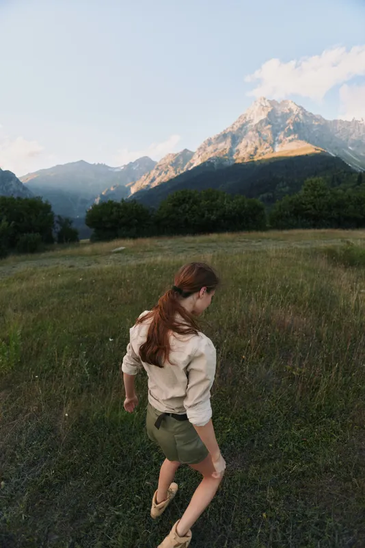 Young woman with long hair walks through a grassy meadow toward distant mountains, capturing a calm lifestyle moment outdoors and feeling free