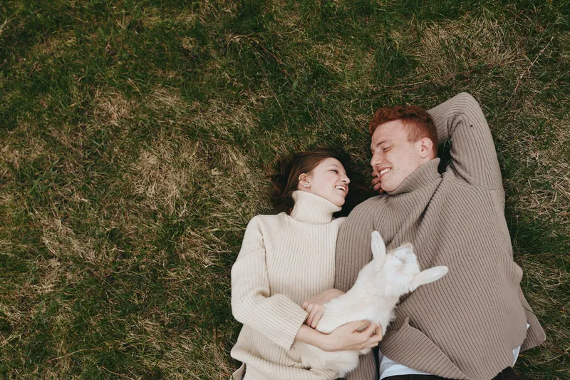Young couple lies on grass holding a baby goat while smiling and enjoying a cozy autumn day in warm knitted sweaters outdoors.