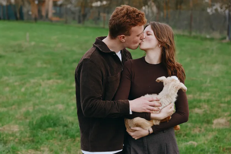 Joyful couple outdoors kissing while holding a curious goat kid, sunlit meadow scene, casual outfits, relaxed mood, natural greens, rustic charm, wholesome lifestyle moment