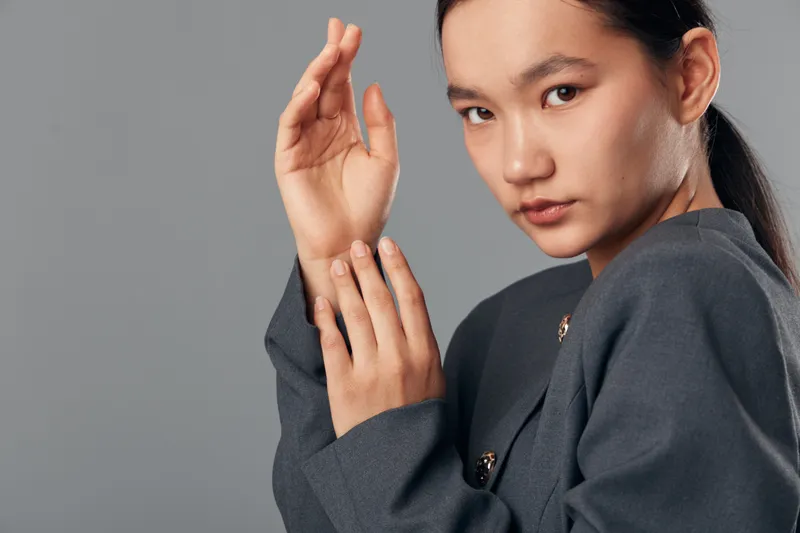 Woman with serious expression wearing stylish gray jacket posing with crossed hands near face against plain gray background for fashion portrait and contemporary style.