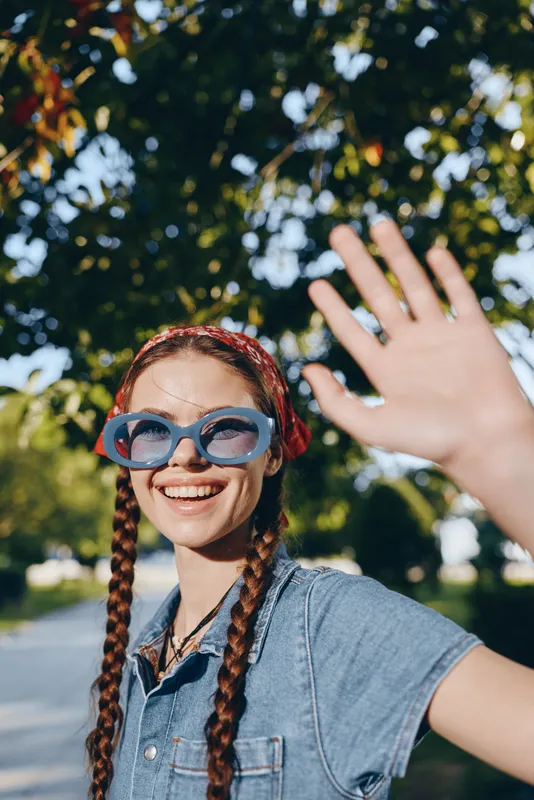 smiling young woman with braids wearing blue sunglasses and denim shirt waving hand outdoors under sunny sky with green trees background