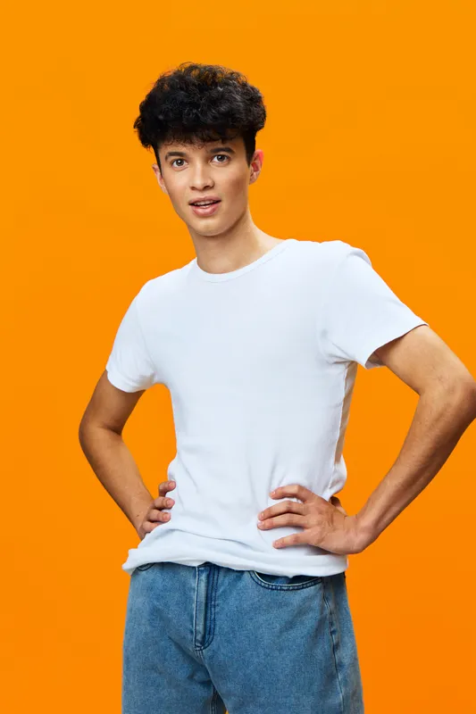 young man standing confidently with hands on hips wearing white t-shirt and blue jeans against bright orange background, casual style, curly black hair