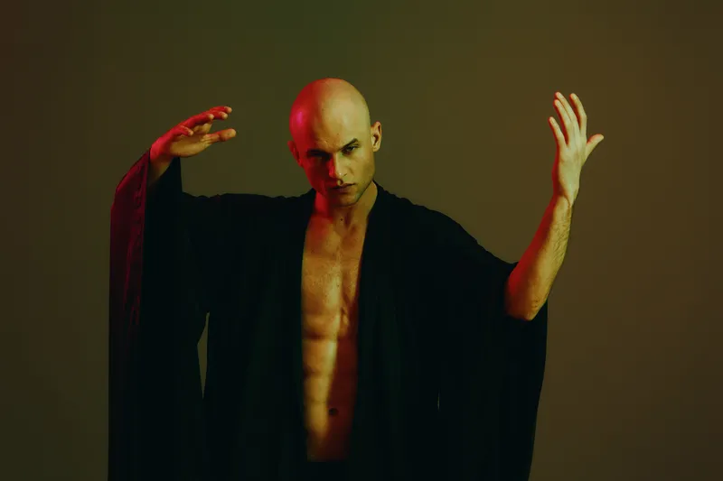 A striking man in a black robe poses dramatically with expressive hand gestures, creating an intriguing atmosphere with his intense look and moody background.