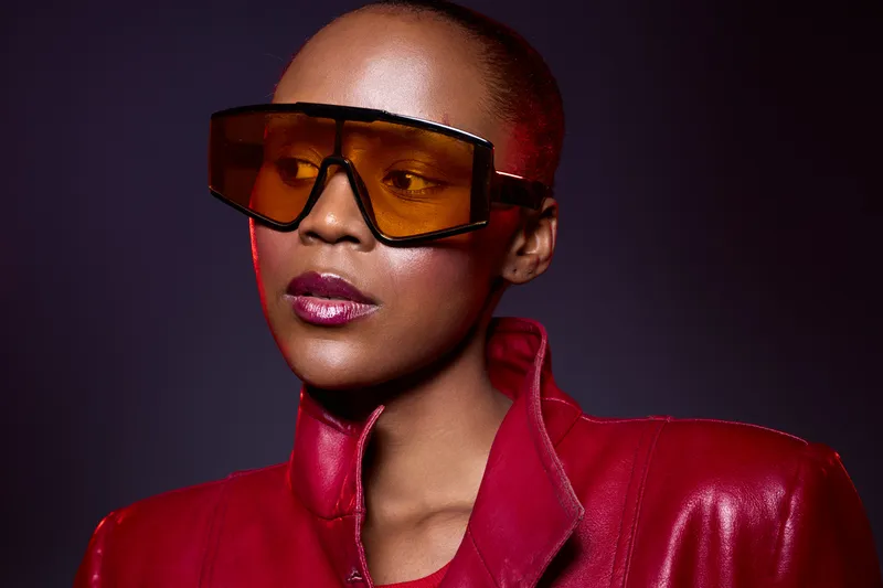 Stylish dark skinned African woman wearing oversized orange sunglasses and a red leather jacket looks away on a dark purple background. Concept of fashion, confidence, and modern style.
