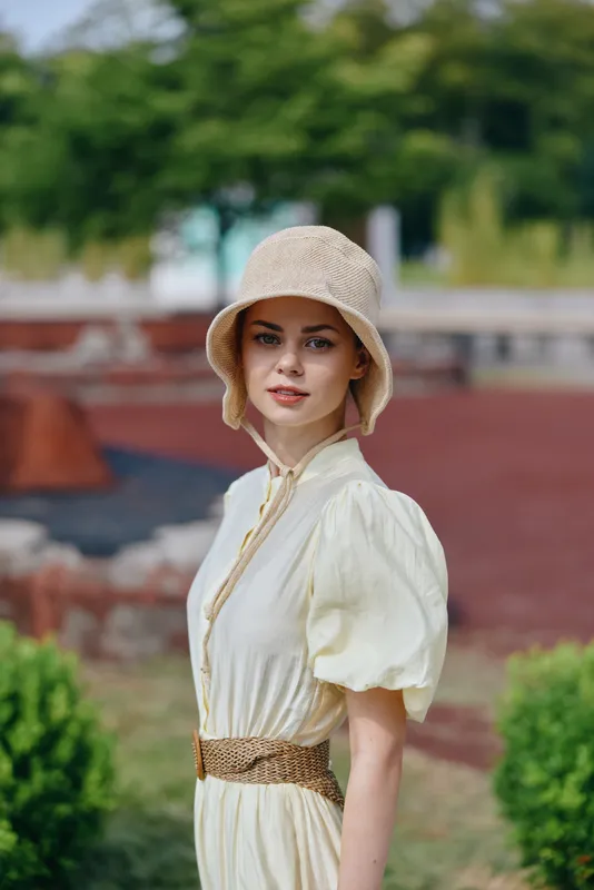 Vintage woman in a cream dress and bucket hat poses outdoors in a garden, looking over shoulder with a calm, confident expression, conveying timeless fashion