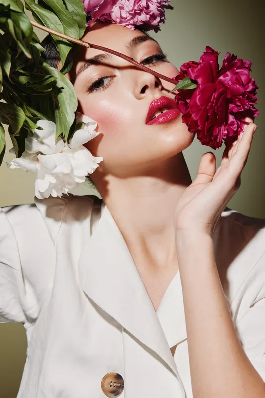 Close-up of a young woman with glossy red lips and dewy makeup holding vibrant pink and white flowers near her face. Studio lighting enhances her flawless skin and fashion elegance.