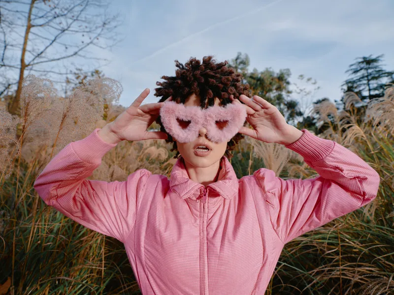 Woman wearing a pink hoodie and fuzzy mask in an outdoor field, a playful fashion portrait amid tall grasses and clear sky, vivid colors and creative street style.