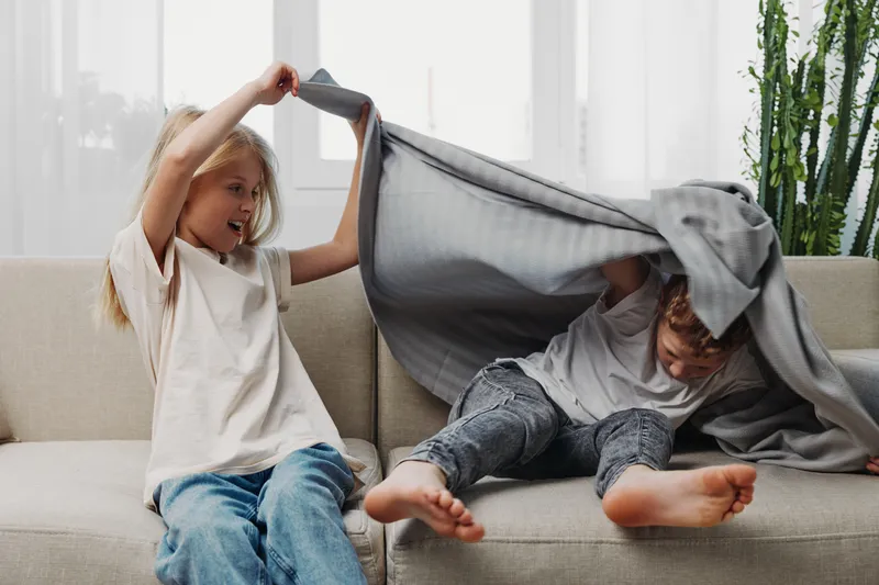 Two playful children on sofa having fun with gray blanket in bright living room, showing joy and sibling interaction during indoor leisure time.
