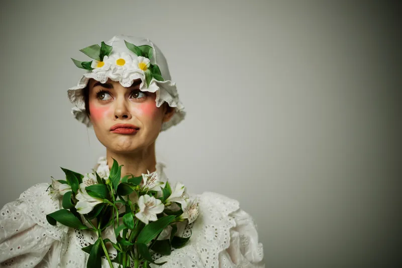 Female model with flowers in a whimsical white dress, expressing playful emotions and vintage style in a soft, neutral background