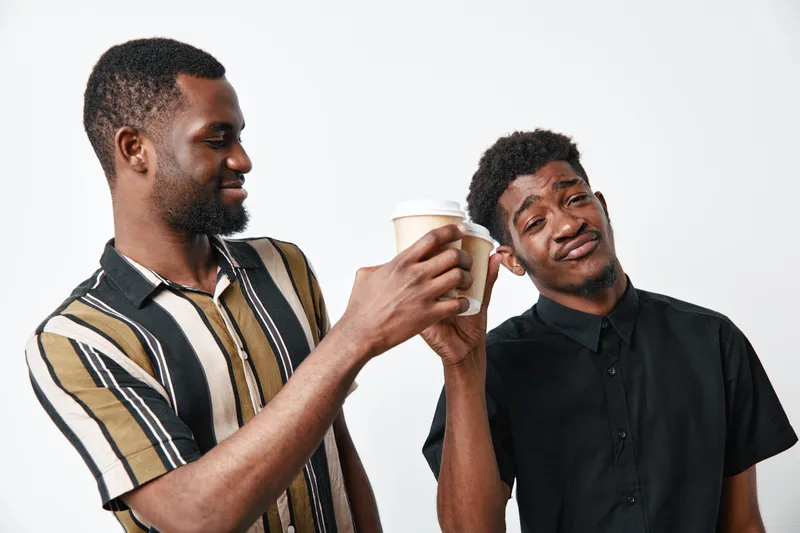 Two African American men with dark skin clinking disposable coffee cups in celebration, casual clothing and friendly gesture on white background studio.