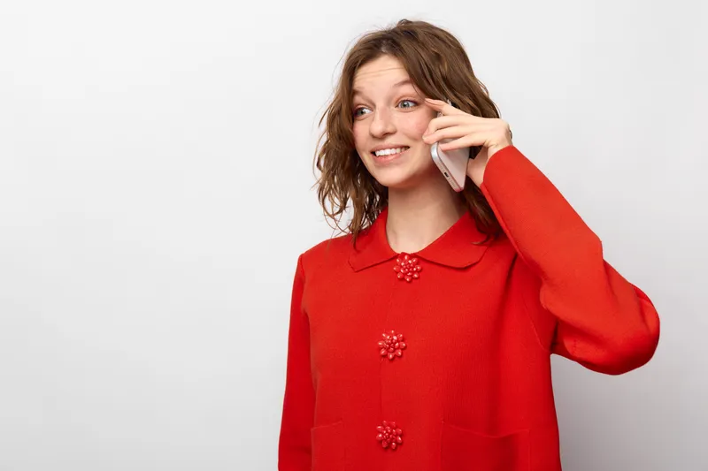 Smiling young woman in red sweater talking on smartphone, looking away with happy expression. Isolated on white background. Concept of communication, technology, and positive mood.