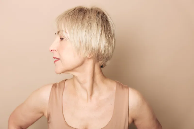 Profile of a mature woman with short blonde hair wearing a beige sleeveless top on a soft beige background. Concept of aging beauty, confidence, and natural lifestyle.