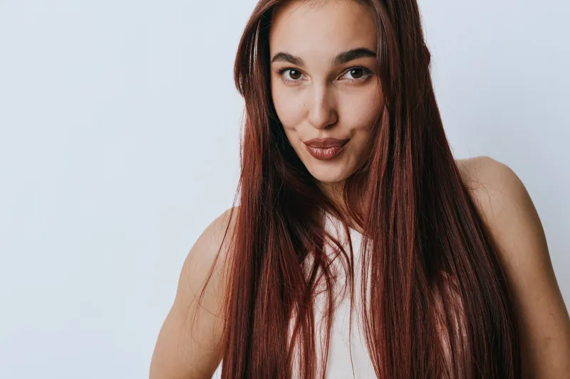 Young woman with long straight hair posing and looking at camera with playful expression on light background. Female model with natural makeup and casual style in studio portrait.