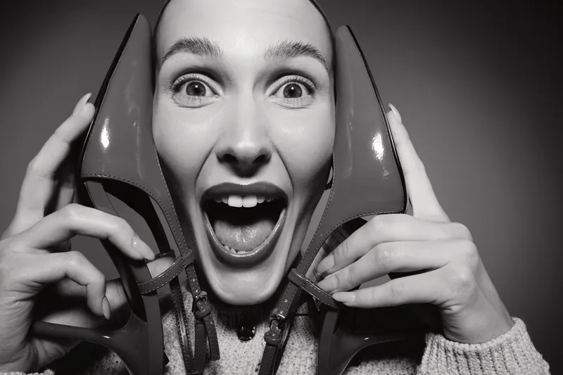 Excited Caucasian woman with light skin holding red high heels near face, wide open mouth and eyes, studio portrait in black and white. Concept for fashion and joy.