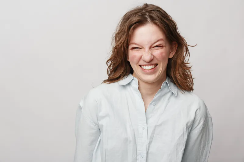 Happy young woman with curly hair smiles broadly wearing white shirt on light background. Portrait of cheerful female with natural expression and casual style in studio lighting.