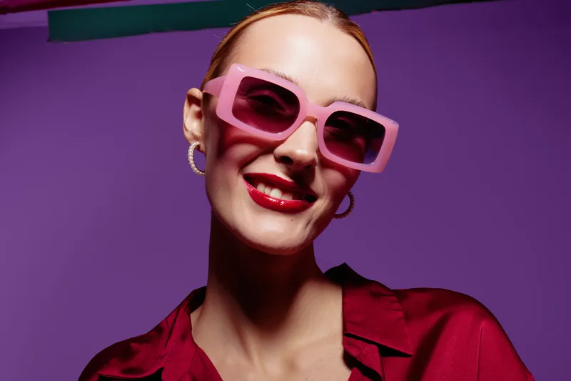 Smiling woman with pink sunglasses and red lipstick poses against purple background. Fashion portrait with stylish accessories and confident expression. Concept of beauty and modern style.