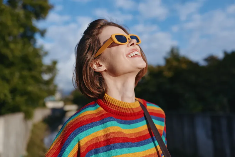 Young adult woman smiling, looking up while wearing yellow sunglasses and rainbow striped sweater outdoors in sunny urban park; colorful fashion, joyful travel lifestyle portrait.