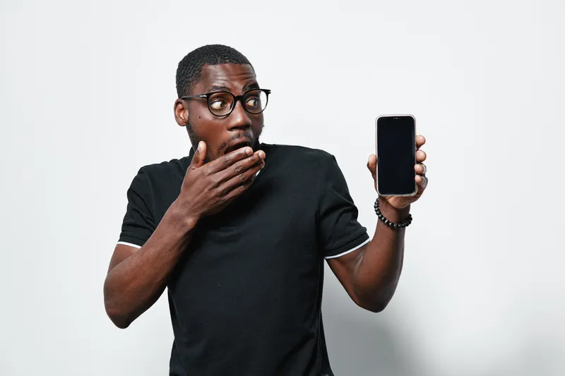 Black man with dark skin and glasses showing smartphone with blank screen in hand, surprised expression and casual black t shirt on white background for technology concept.