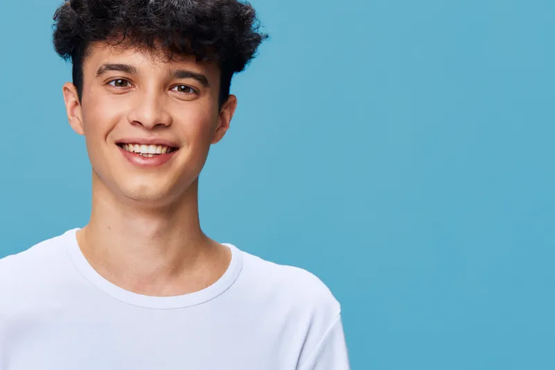 Smiling man with curly hair wearing white t-shirt on blue background, cheerful expression, casual style, studio portrait, youthful and relaxed vibe.