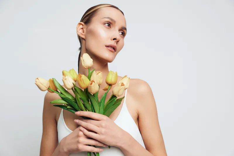 Woman portrait in studio, holding yellow tulips and flowers, spring beauty with calm gaze, white background, soft light, upright posture, fresh touch minimal composition