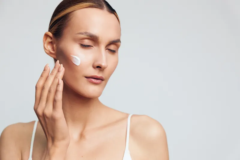 Woman with closed eyes applying white face cream on cheek. Skincare routine concept with natural light and neutral background. Focus on healthy skin and self-care.