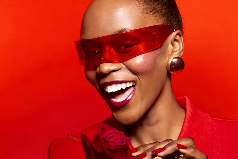 Smiling black African woman with dark skin wearing red sunglasses and earrings poses on red background. Fashion concept with bright makeup, confident expression, and stylish accessories.