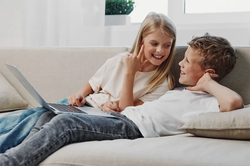Happy children relax on sofa using laptop, smiling and interacting in bright living room. Concept of family time, technology, learning, and sibling bonding indoors.
