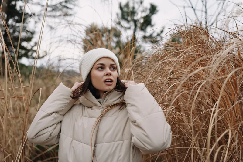 Winter scene with a woman in a light coat among tall beige grasses, wearing a knit hat, looking away, capturing calm outdoor weather and natural texture.