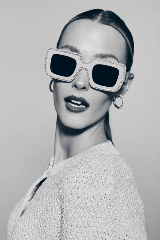 Stylish white woman with pale skin wearing large square sunglasses and hoop earrings poses in knitted sweater. Fashion portrait in black and white studio setting.