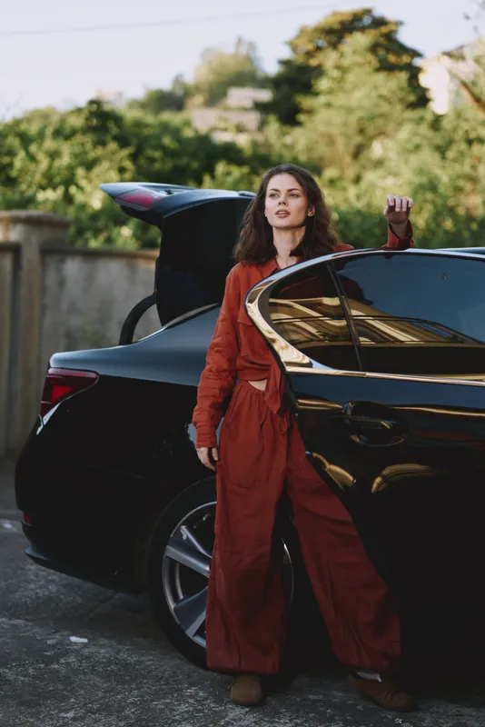 Woman outdoors beside a sleek car, wearing a red outfit, relaxed pose, natural lighting, candid lifestyle moment, urban greenery in the background, confident expression