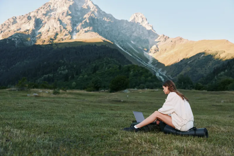 A lone remote worker sits on a grassy meadow with a laptop, surrounded by a mountain landscape. The tranquil scene conveys focus, creativity, and productive momentum in nature.