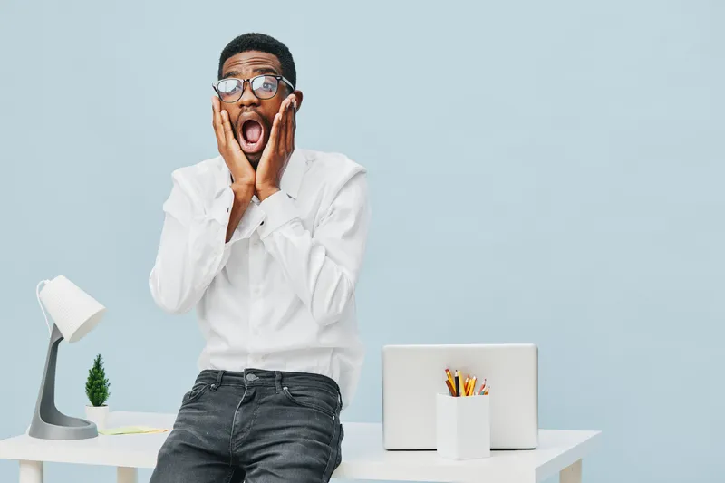 Frustrated young man with glasses wearing white shirt sitting at modern workspace with laptop and stationery, expressing shock or surprise, isolated on light blue background. People emotion concept