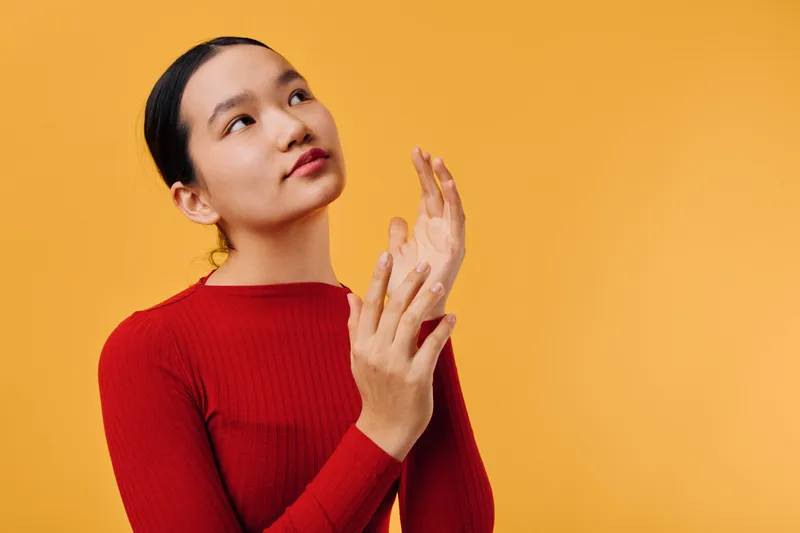 Young woman in red sweater looking up with thoughtful expression on orange background. Asian female model with smooth skin and dark hair posing in studio with natural light.