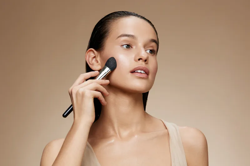 A young woman applying makeup with a brush on her cheek, showcasing a natural look, perfect for beauty and skincare concepts in a soft, neutral background