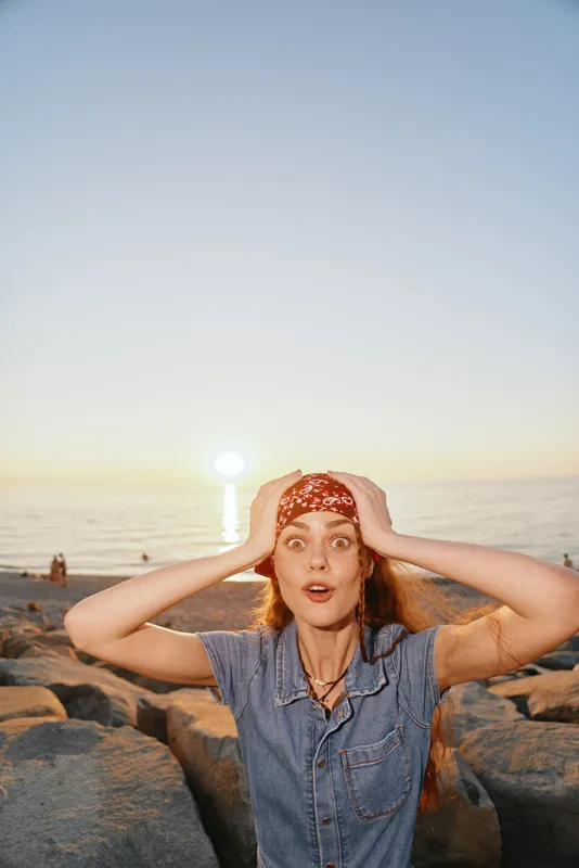 Lifestyle woman in modern boho-western denim street style with film color effect posing surprised at sunset near rocky beach