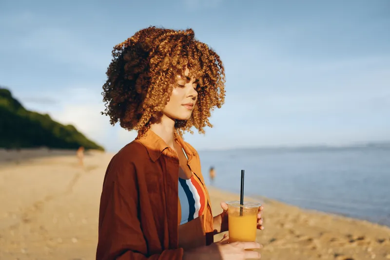 Woman with curly hair stands on the beach by the ocean, holding a cold drink with straw, summer outdoors portrait, sunlight golden light, calm relaxed gaze