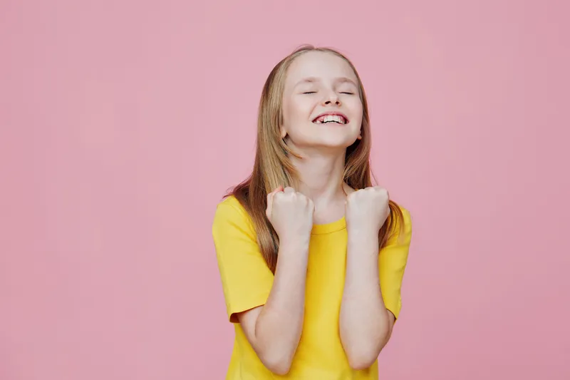 Cheerful girl with long hair celebrating success in a yellow t shirt against a pink background, expressing happiness and joy