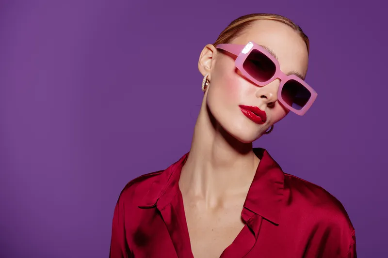 Stylish woman with pink sunglasses and red lipstick poses against purple background. Fashion portrait with vibrant colors and confident expression. Concept of beauty and modern style.