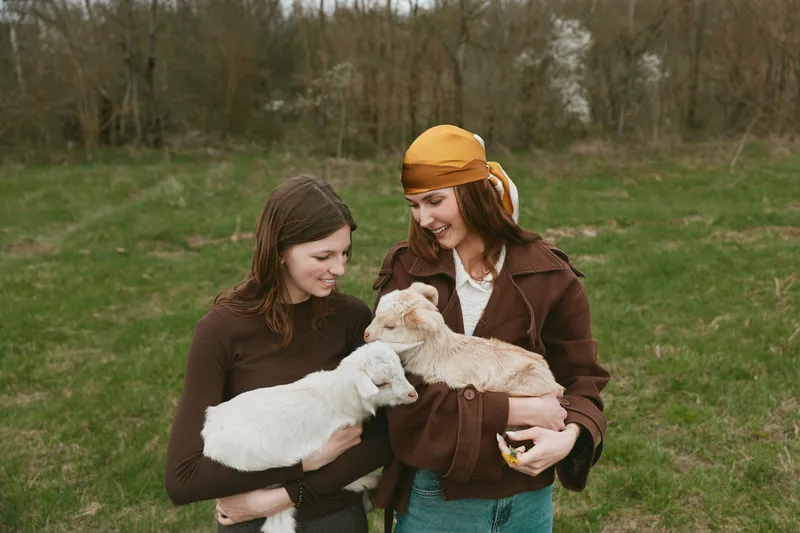 Two young women enjoy a spring day outdoors holding baby goats in a green meadow, smiling and connecting with nature in casual warm clothing.