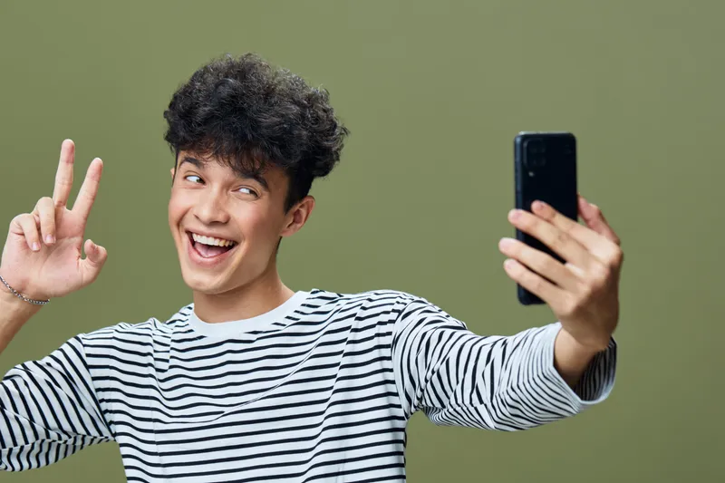 young man taking selfie smiling and showing peace sign with striped shirt and curly hair on green background, happy expression, casual style portrait