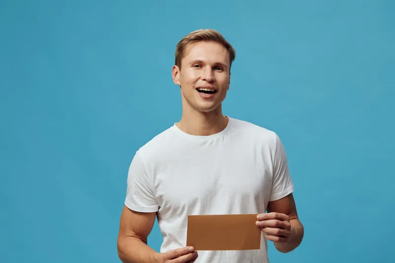 smiling young caucasian man hold brown envelope wearing white t-shirt isolated on blue background digital detox concept