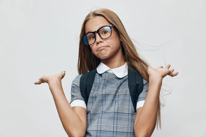 Girl child with glasses and long hair wearing school uniform and backpack shrugs shoulders and shows confusion or uncertainty on gray background for education concept.