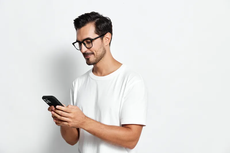 Smiling man in casual white t-shirt holds smartphone, standing against plain solid background. Modern lifestyle and technology concept.