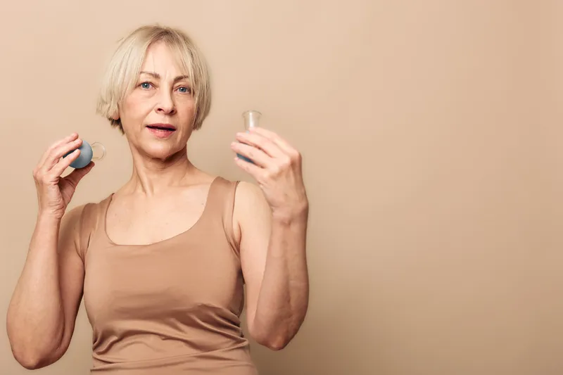 Mature woman with short blonde hair holds two cosmetic containers in beige tank top against neutral background. Concept of skincare routine, beauty care, and self-care rituals.