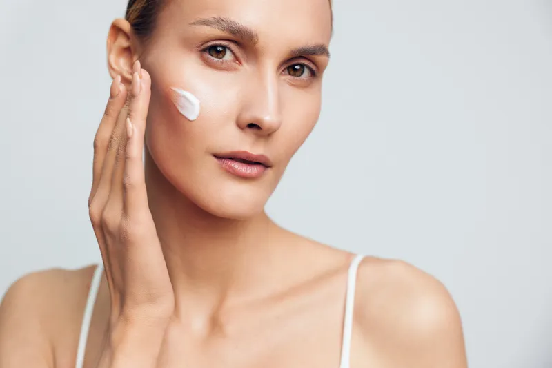 Young woman applies face cream on cheek with hand closeup natural skin care beauty routine isolated on light background with soft natural light and calm expression.