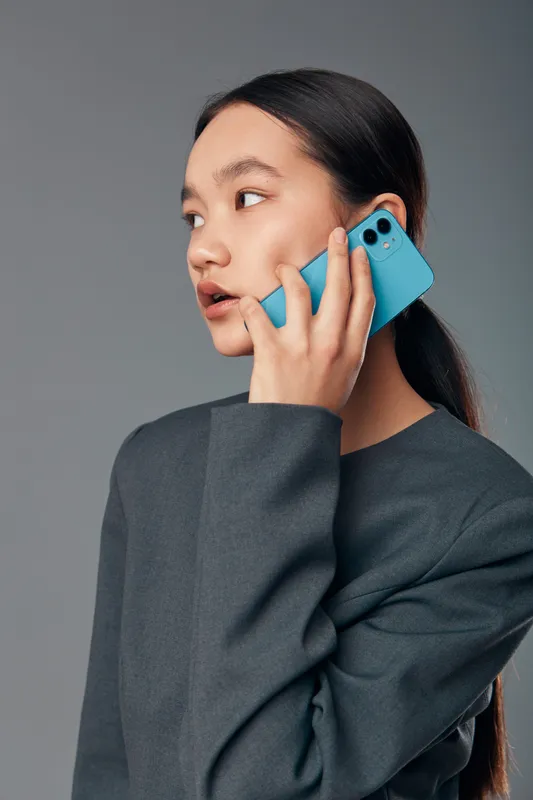 Young woman with long black hair and gray sweater talking on turquoise smartphone. Portrait of thoughtful female communicating on mobile phone with gray background. Side view closeup.